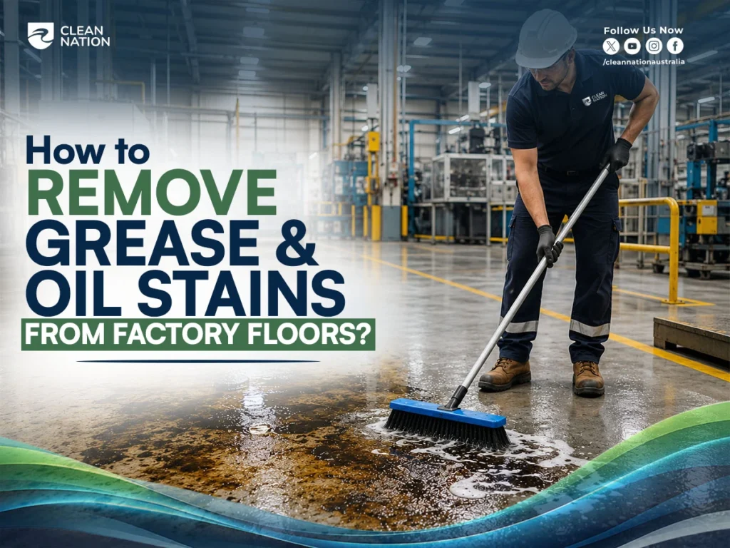 A Clean Nation worker scrubs grease and oil from a large industrial factory floor using a push broom, demonstrating How to Remove Grease and Oil Stains From Factory Floors? in a clean, professional manufacturing environment.